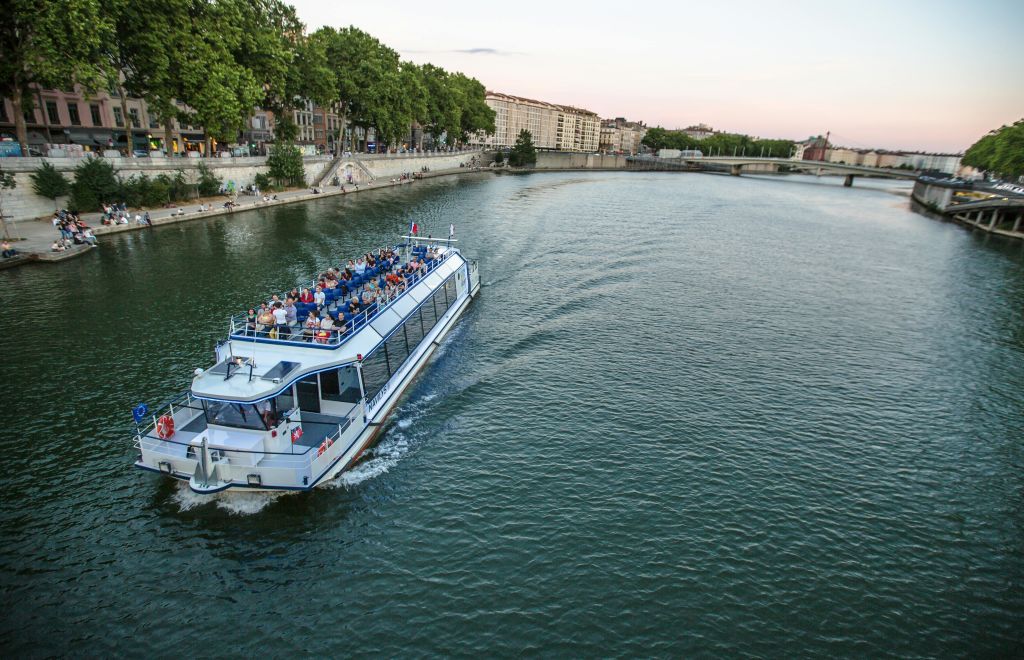 나무와 건물이 늘어선 강둑 근처의 강에서 승객과 함께 항해하는 보트.