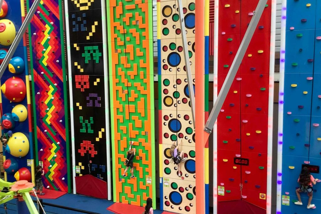 다양한 기하학적 패턴과 한쪽 벽에 타이머가 보이는 다채로운 실내 등반 벽을 등반하는 두 사람.