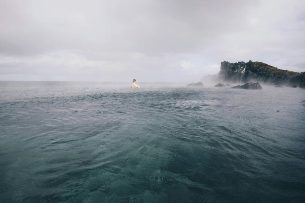 바위 절벽 근처의 바다에 서서 흐린 하늘 아래 물에서 안개가 솟아오르는 사람.