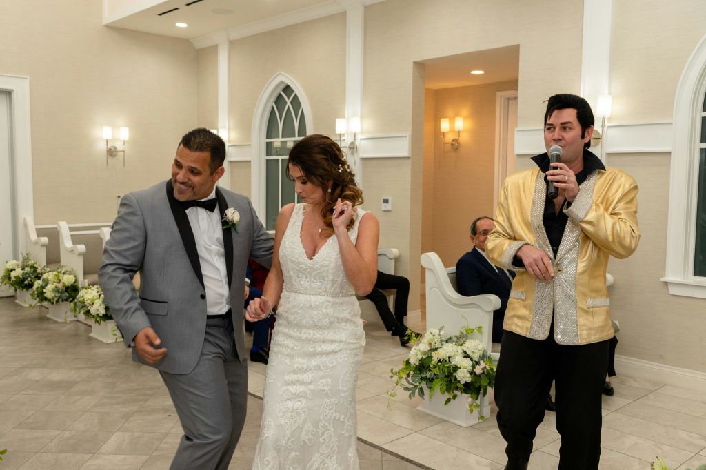 엘비스의 옷을 입은 남자가 마이크를 잡고 노래하는 결혼식에서 신랑 신부가 춤을 추고 있습니다.