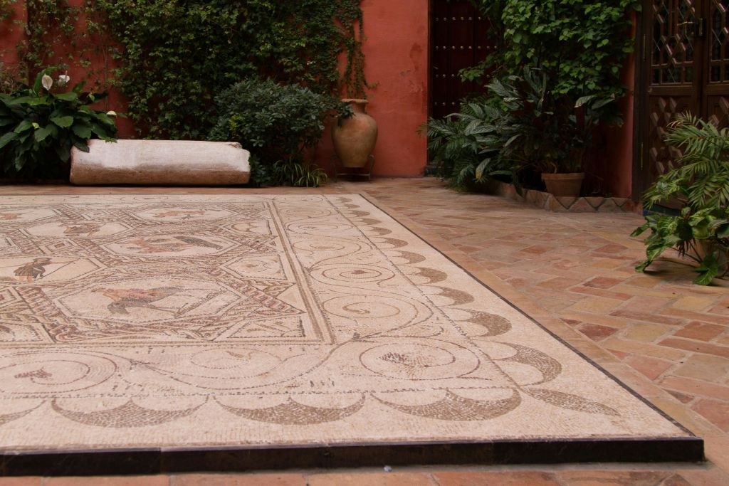 울창한 녹음과 화분, 항아리가 있는 붉은 벽으로 둘러싸인 디테일한 모자이크 바닥이 있는 마당입니다.