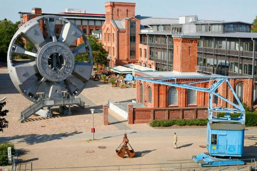 산업 유산 건물, 커다란 금속 바퀴 구조, 파란색 크레인, 걷는 사람이 있는 마당입니다.