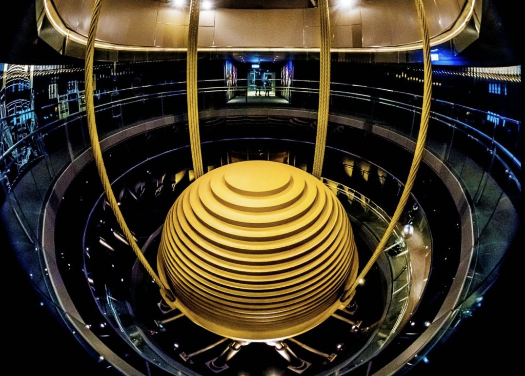 원형의 다층 실내 공간에 케이블로 매달린 크고 노란색 구형 구조. 희미한 조명.