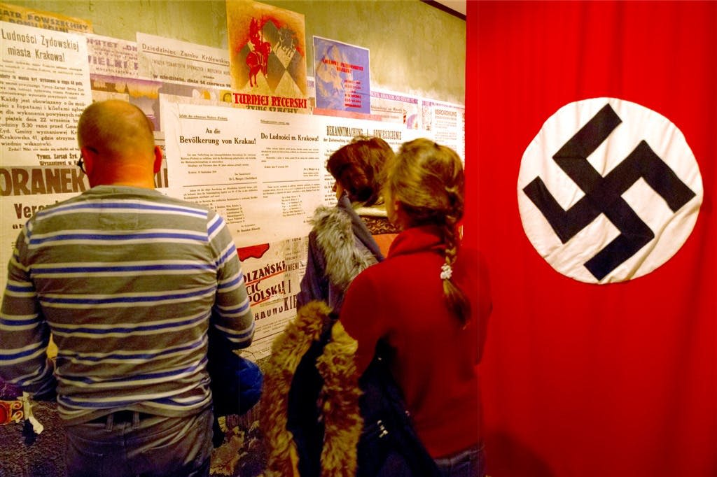 흰색 동그라미에 검은색 스와스티카가 그려진 커다란 붉은 깃발 옆 벽에 걸린 역사 포스터를 읽는 사람들.
