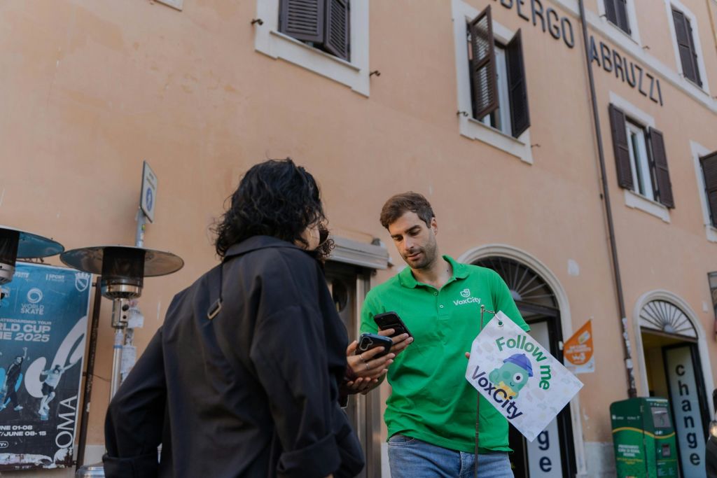 간판을 들고 있는 초록색 "VoxCity" 셔츠를 입은 사람이 아치형 창문이 있는 건물 근처에서 다른 사람과 이야기하고 있습니다.