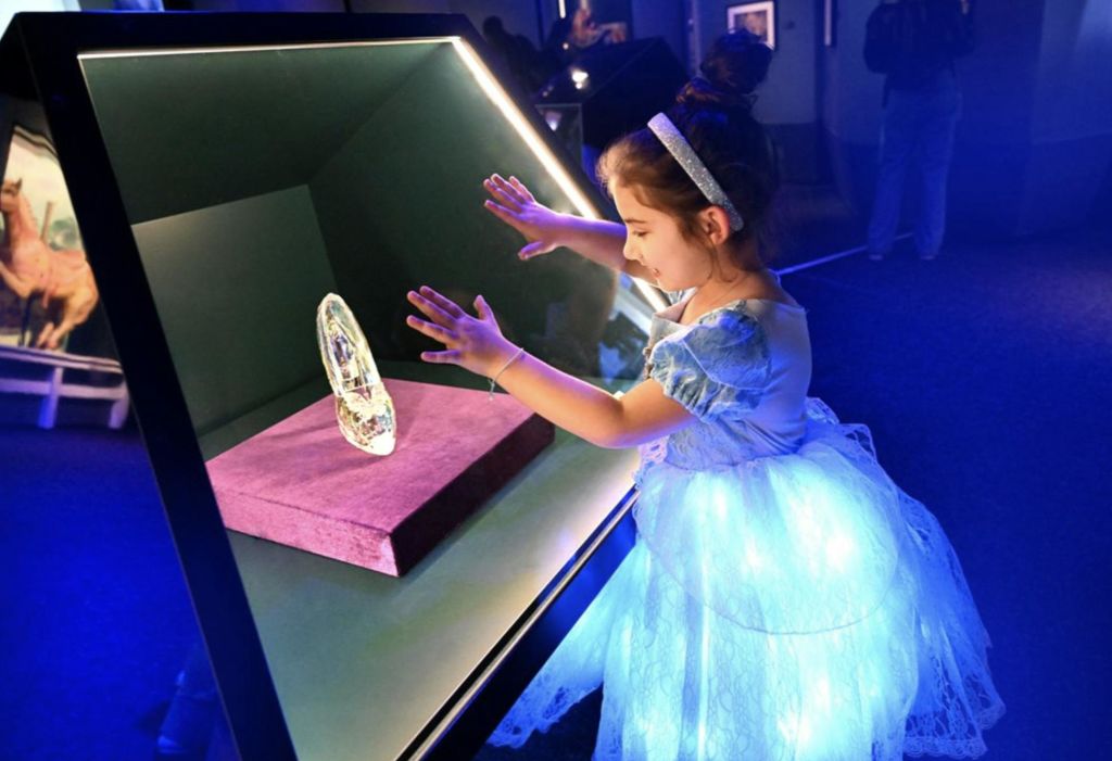 반짝이는 파란색 드레스를 입은 어린 소녀가 전시회에서 유리 디스플레이 내부의 크리스탈 유물을 바라보고 있습니다.