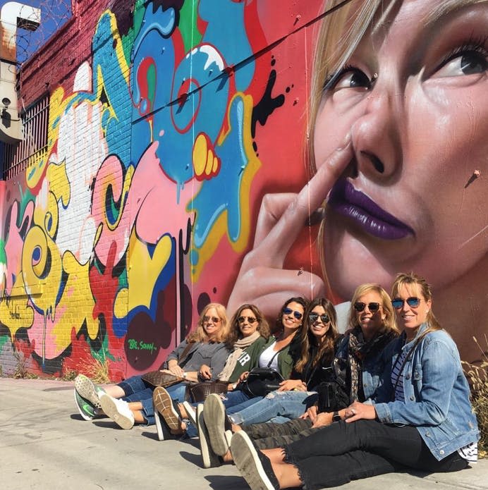 커다란 얼굴 벽화가 그려진 화려한 그라피티 벽 앞 보도에 앉아 있는 한 무리의 여성들.