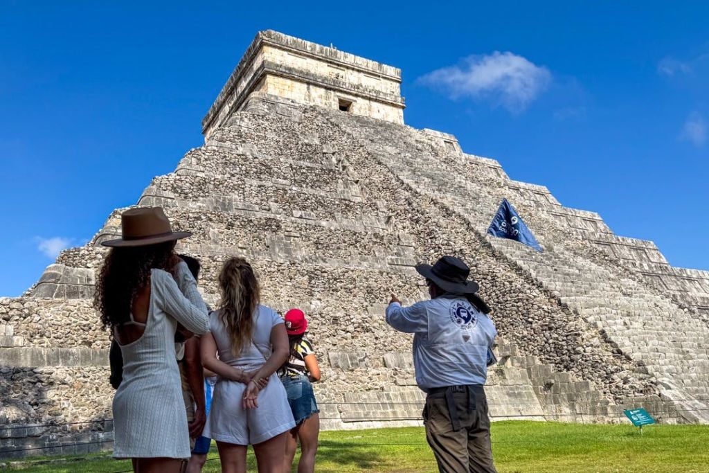 한 무리의 사람들이 맑고 푸른 하늘 아래 크고 오래된 석조 피라미드 앞에서 가이드의 말을 듣고 있습니다.