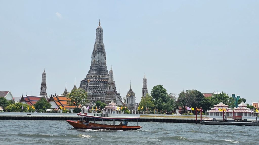 맑은 하늘 아래 커다란 사원 왓 아룬 (Wat Arun) 이 있는 강가의 전통 목조 보트입니다.