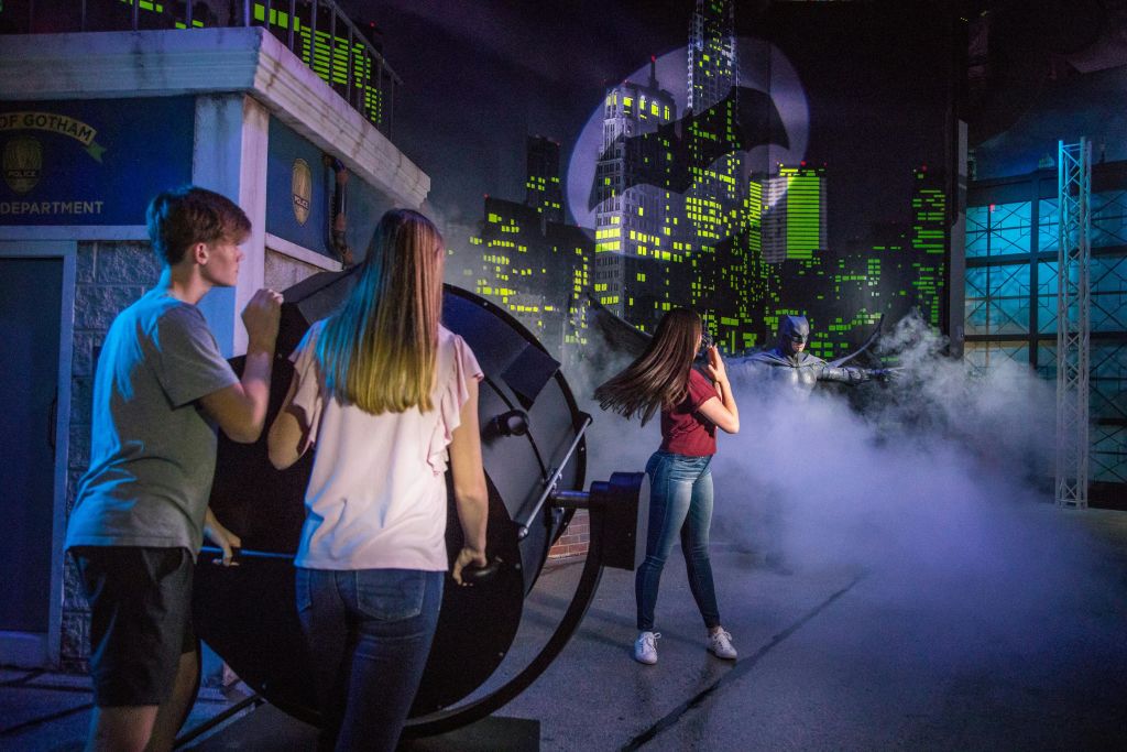 세 사람이 도시 경관을 배경으로 배트맨 심볼이 투영된 대형 스포트라이트를 운영합니다.