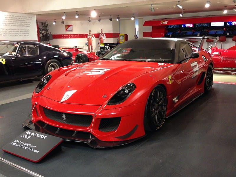 빨간색 페라리 스포츠카가 실내에 전시되어 있으며, 다른 자동차와 정보 표지판으로 둘러싸여 있습니다.