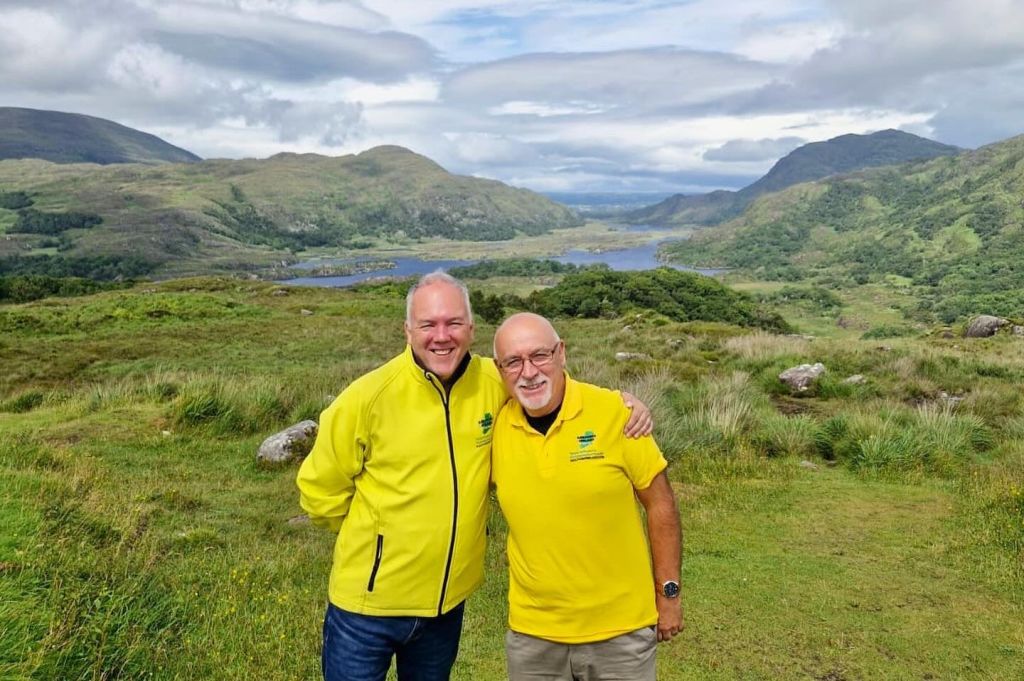 푸른 언덕과 호수를 배경으로 아름다운 경치를 감상하며 노란색 상의를 입은 두 사람이 미소를 짓고 있습니다.