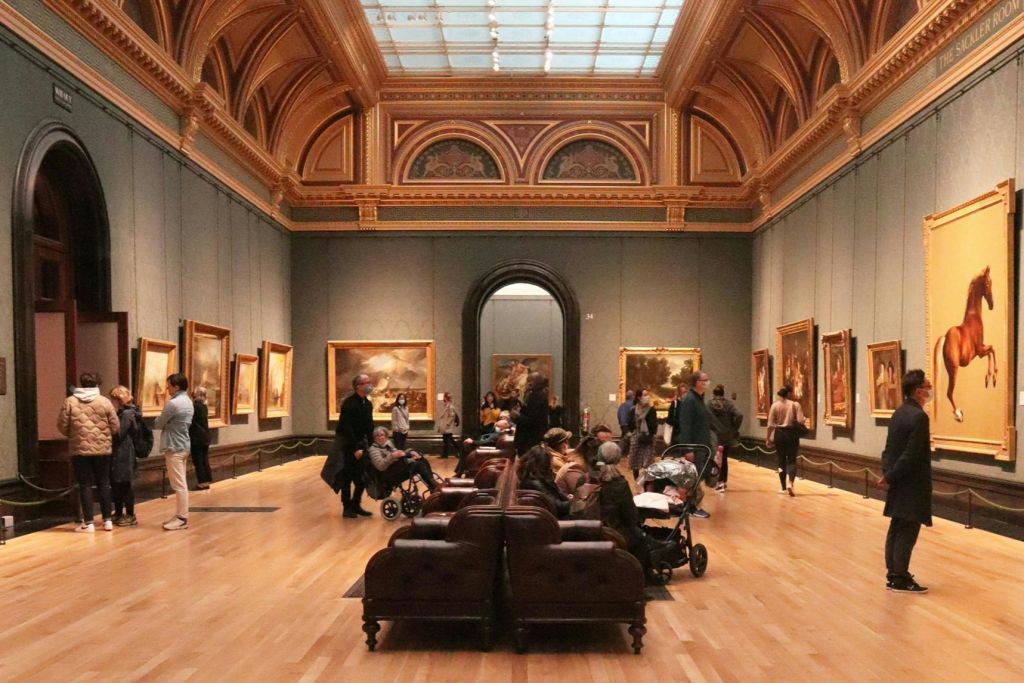 사람들은 중앙에 높고 화려한 천장과 쿠션이 있는 벤치가 있는 넓은 미술관에서 그림을 관찰합니다.