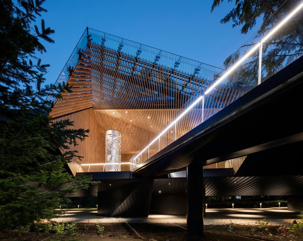 목조 슬랫 외관과 높은 통로를 갖춘 현대적인 건물로 밤에는 조명이 켜집니다. 나무와 커다란 조각 기둥이 있습니다.