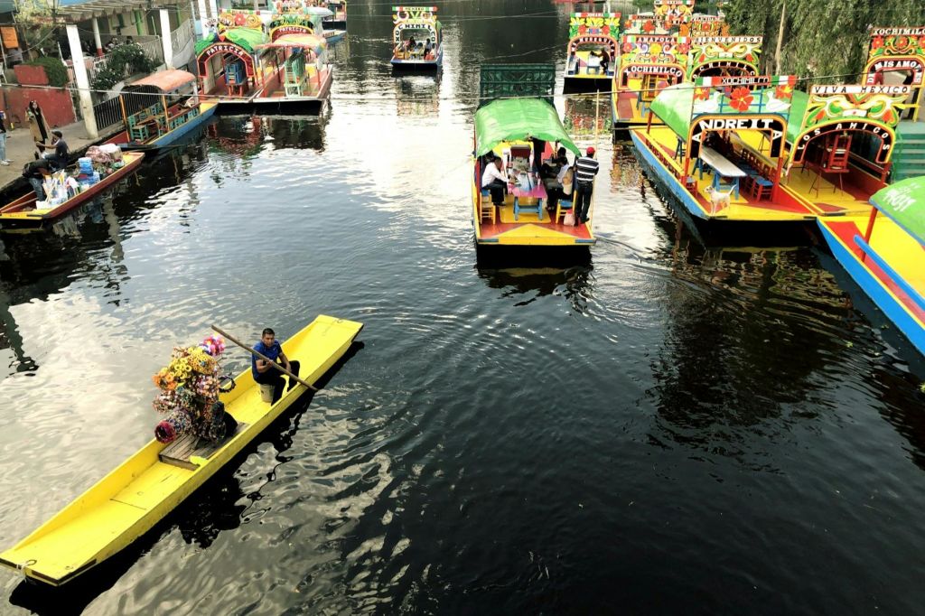 운하에 장식된 캐노피가 있는 밝은 색의 보트. 한 남자가 노란 뗏목을 타고 노를 젓고 있는 두 척의 보트에서 사람들이 보입니다.
