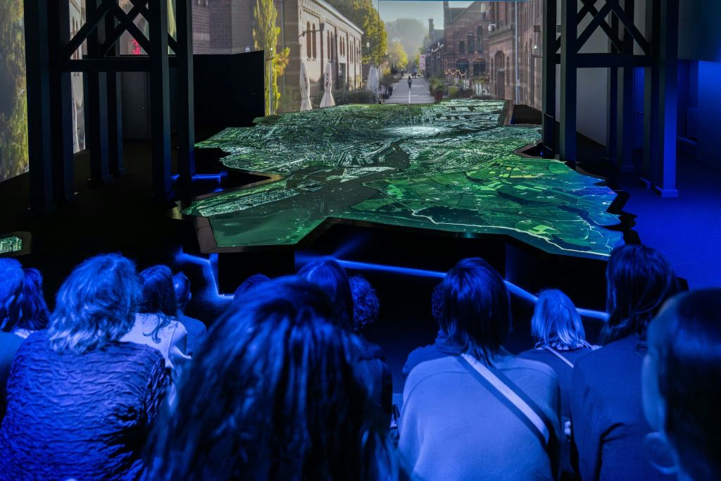 도시 풍경을 배경으로 조명이 어두운 방에서 조명이 켜진 지형도 앞에 관객이 앉아 있습니다.