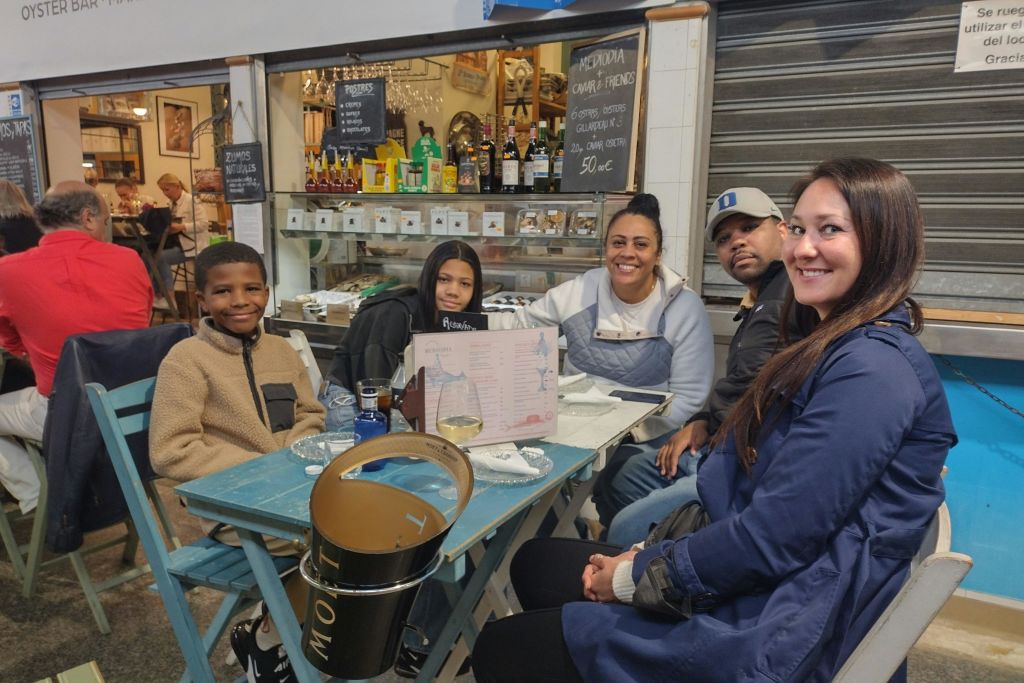 캐주얼한 분위기의 테이블에 네 명이 앉아 카메라를 바라보며 미소를 짓고 있으며, 테이블에는 음료와 메뉴가 놓여 있습니다.