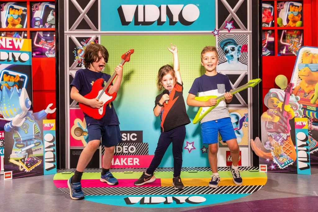 세 명의 어린이가 "VIDIYO" 라는 단어와 함께 다채로운 배경 앞에서 장난감 기타와 마이크를 들고 포즈를 취하고 있습니다.