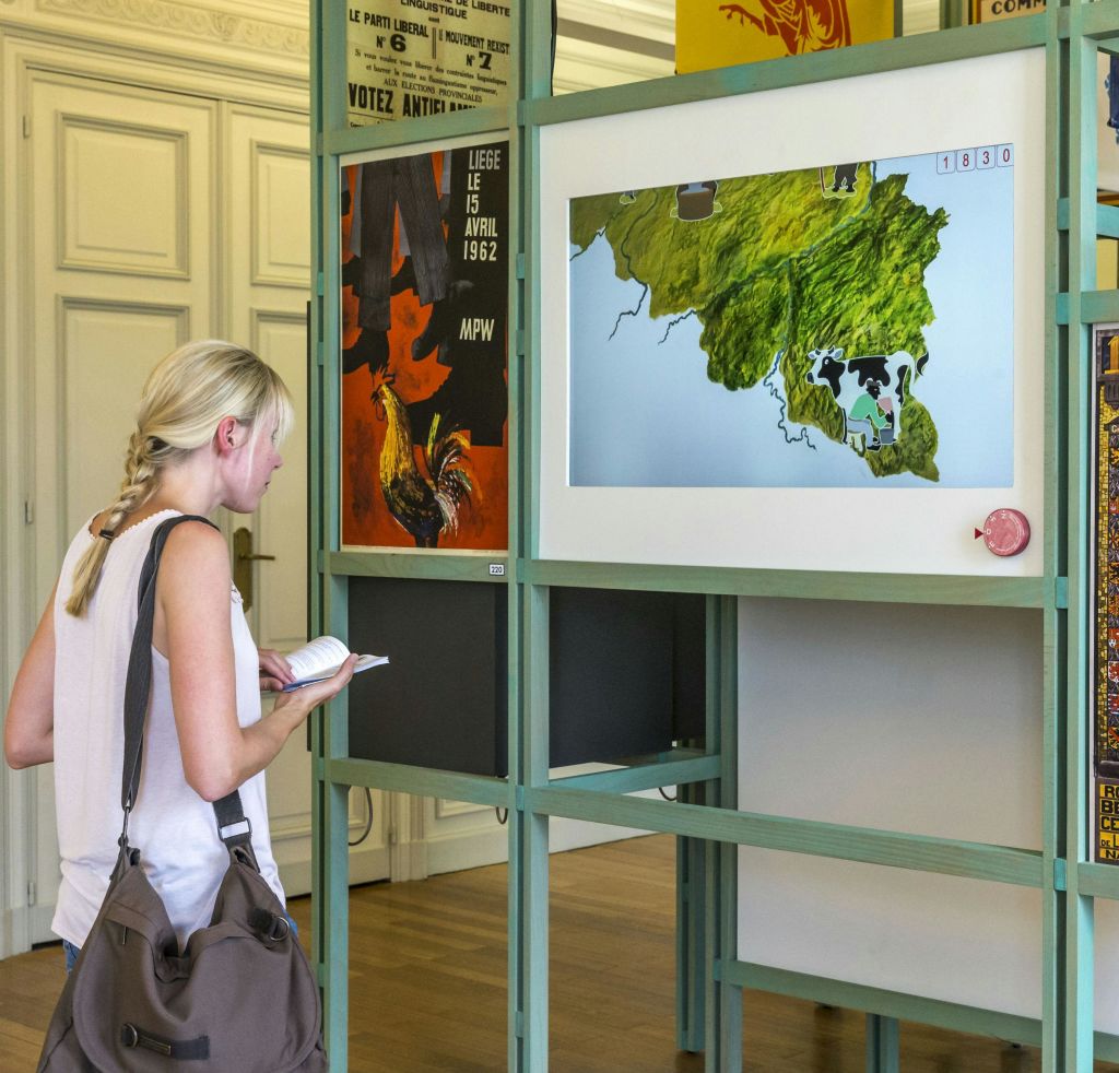 금발의 여성이 역사적인 포스터를 배경으로 지도를 보고 있습니다. 그녀는 메모장을 들고 숄더백을 들고 있습니다.