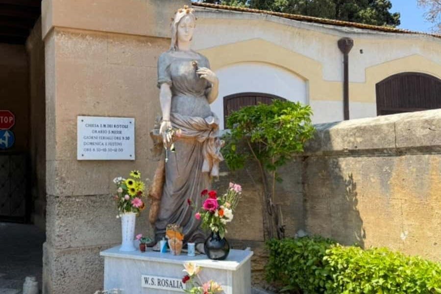 "W.S. Rosalia" 라고 새겨진 받침대에 다양한 꽃꽂이와 제물로 둘러싸인 꽃을 들고 있는 여성의 동상.