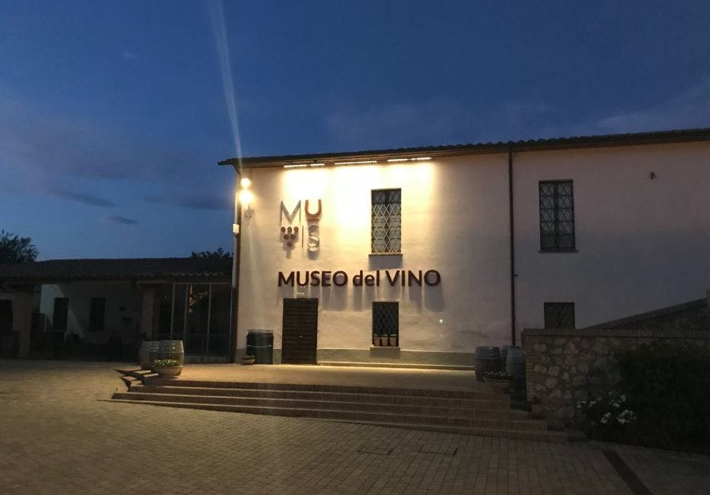 해질 무렵 입구와 표지판을 비추는 조명이 있는 "Museo del Vino" 건물 외부에는 나무 와인통이 있습니다.