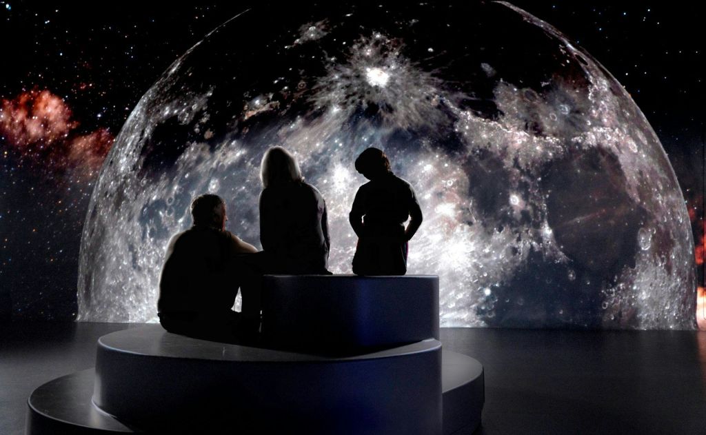 세 사람이 커다랗고 상세한 달 이미지를 배경으로 곡선형 벤치에 앉아 있습니다.