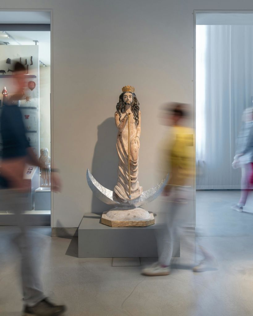 박물관의 받침대에는 왕관을 쓴 인물이 서 있습니다. 흐릿한 형체가 디스플레이 양쪽을 지나갑니다.