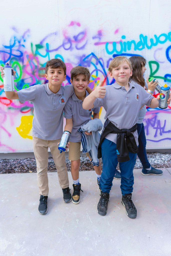 교복을 입은 세 명의 남학생이 낙서로 덮인 벽 앞에서 스프레이 페인트 캔을 들고 웃으며 엄지손가락을 치켜들고 있습니다.