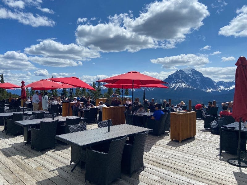 산이 있고 구름이 흩날리는 밝고 푸른 하늘이 있는 빨간 우산 아래 테이블에 앉은 사람들이 있는 야외 식사 공간.