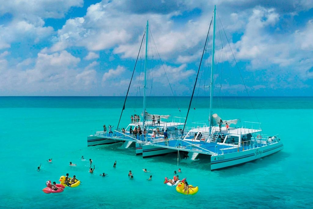 청록색의 맑은 물에 정박한 두 쌍동선은 부분적으로 흐린 하늘 아래에서 사람들이 수영하고 팽창식 뗏목에 떠 있습니다.