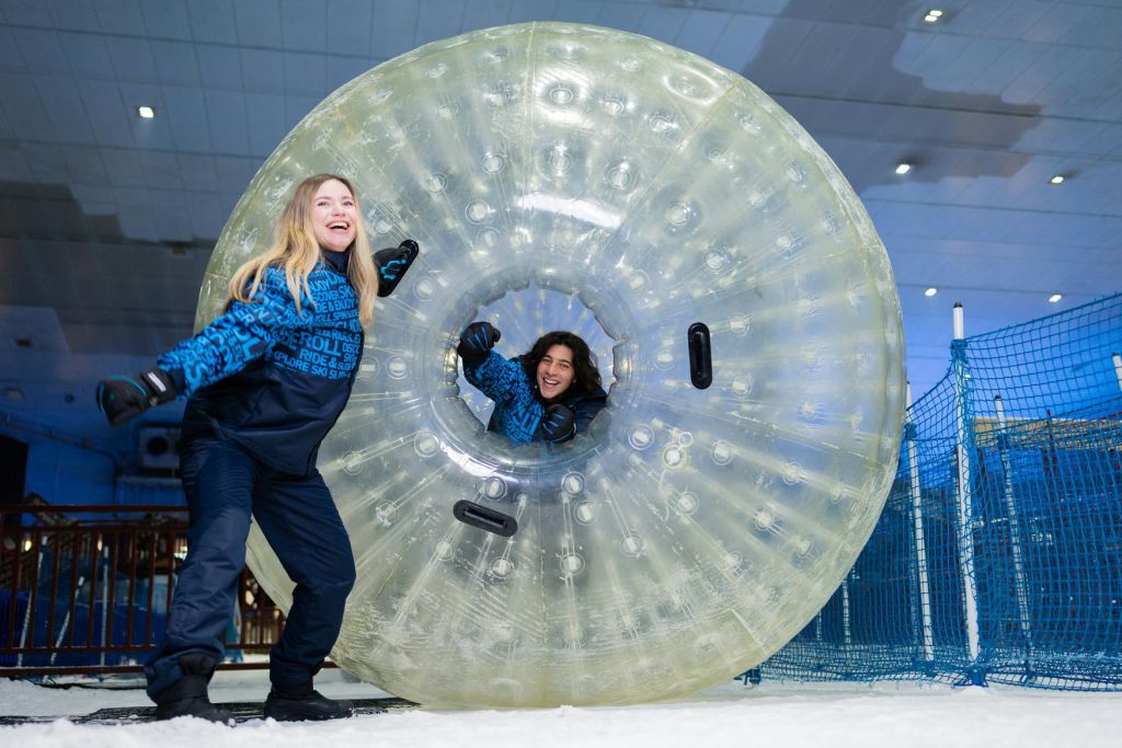 겨울 장비를 입은 두 사람이 실내 스노우 파크를 즐기고 있습니다. 하나는 팽창식 조브 볼 외부에 있고 다른 하나는 내부에 있습니다.