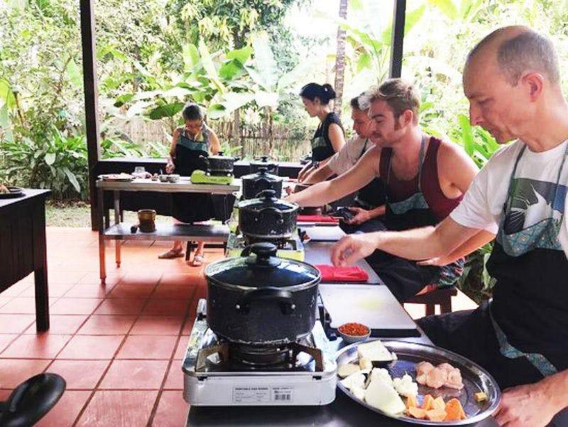 무성한 녹음으로 둘러싸인 야외 스테이션에서 냄비와 프라이팬으로 요리하는 앞치마를 입은 사람들.
