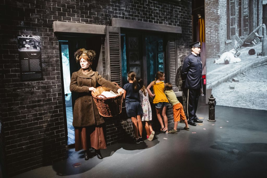역사적인 복장을 한 여성이 벽돌 건물 밖에서 바구니를 들고 있습니다. 아이들은 그녀의 옆에서 창문을 들여다봅니다.