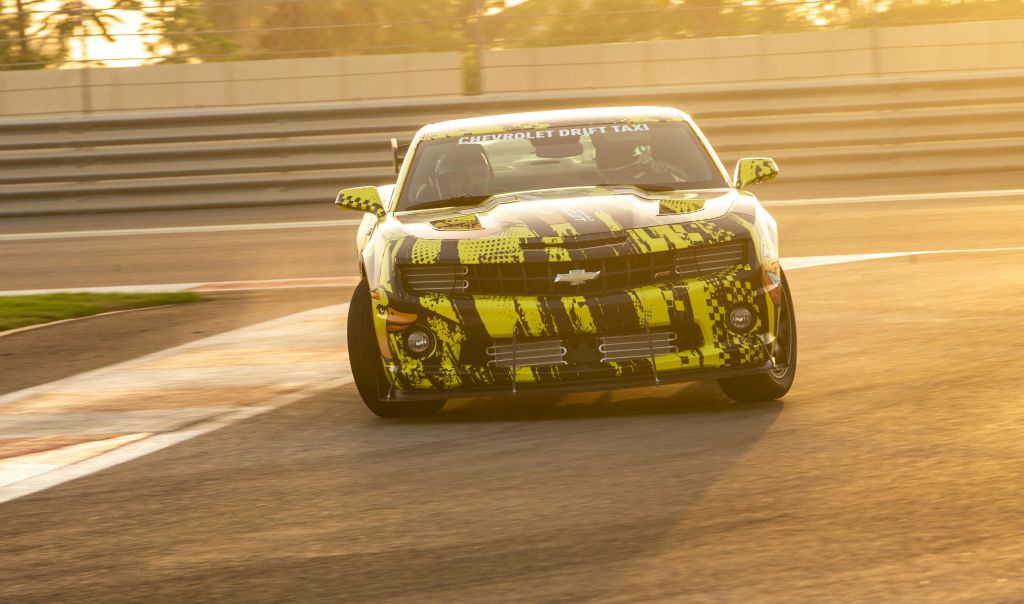 앞유리에 "쉐보레 드리프트 택시" 가 있는 밝게 칠해진 스포츠카는 석양이 질 무렵 레이스 트랙에서 주행합니다.