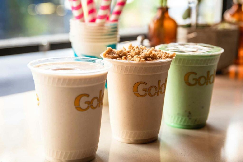 "골드" 라고 적힌 플라스틱 컵에 세 개의 밀크셰이크를 넣고, 하나는 부서진 쿠키를 얹습니다. 분홍색 줄무늬 빨대가 배경에 있습니다.