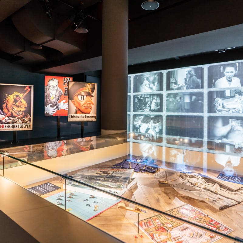 제2차 세계대전 선전 포스터, 유물이 전시된 유리 진열장, 흑백 이미지가 투영된 벽이 있는 박물관 전시물입니다.