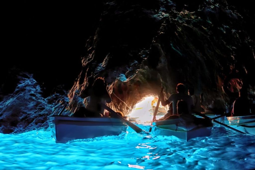 밝은 출구를 향해 빛나는 푸른 동굴 사이로 사람들이 노를 젓고 있는 두 척의 보트. 동굴 벽은 어둡고 바위투성이입니다.