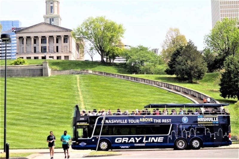 역사적인 건물 앞에 있는 2층 투어 버스와 나무가 있는 잘 다듬어진 잔디밭. 인근을 걷는 두 사람.