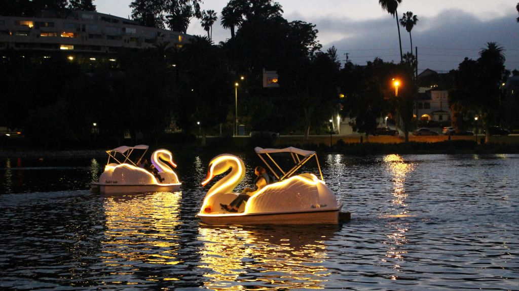 해질 무렵 호숫가에 조명이 켜진 백조 모양의 페달 보트 두 대와 내부의 사람들. 나무와 가로등이 배경으로 보입니다.