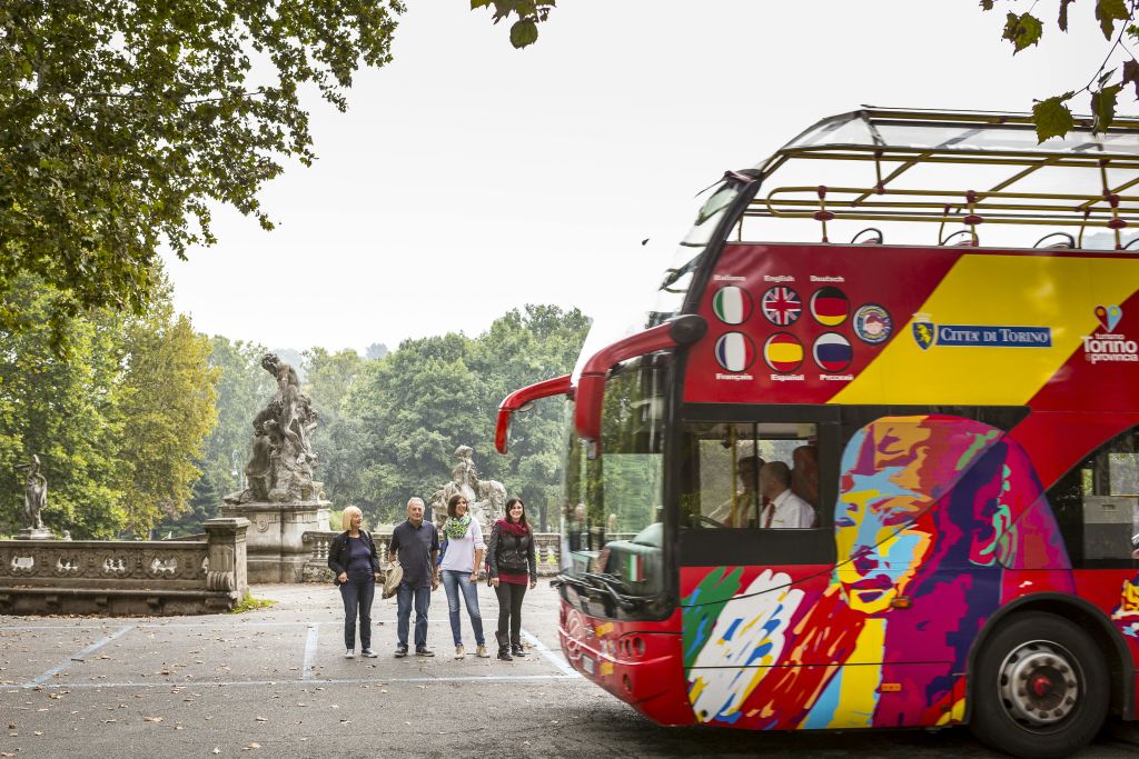 녹색 공원의 동상 옆에 주차된 다양한 깃발로 장식된 화려한 2층 투어 버스 근처에 4명이 서 있습니다.