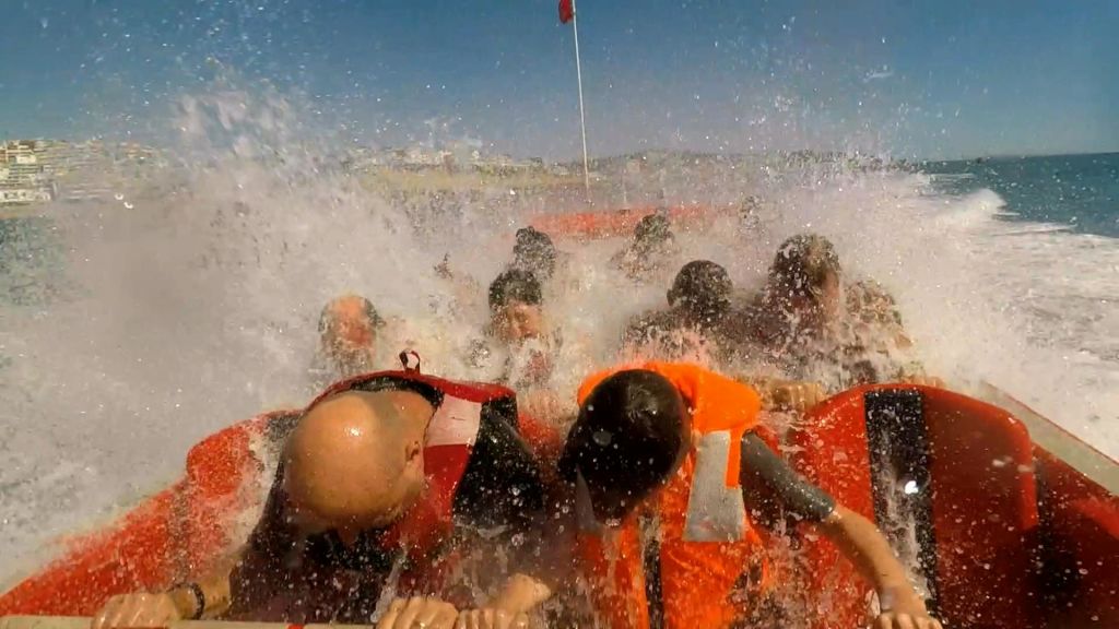 보트에 주황색 구명 조끼를 입은 한 무리의 사람들이 맑고 푸른 하늘 아래에서 커다란 파도에 휩싸이고 있습니다.