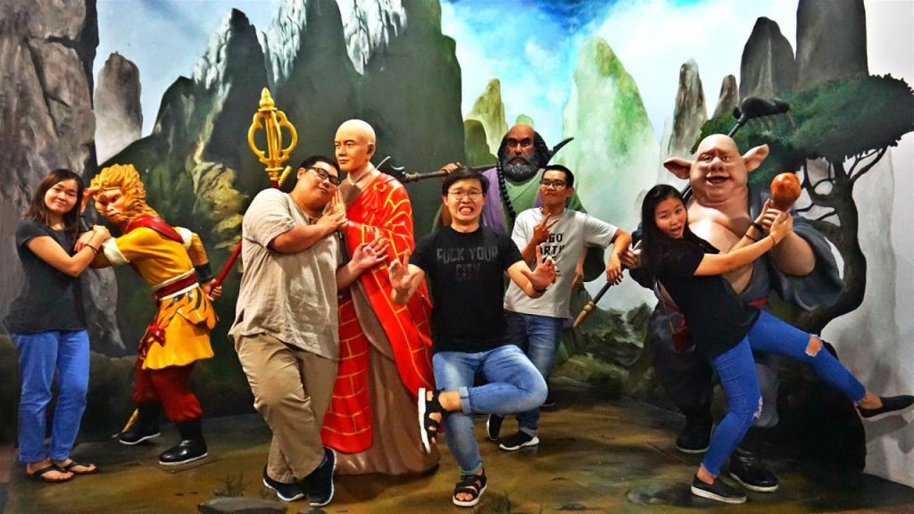 산과 하늘을 배경으로 사실적인 승려상과 신화 속 인물상을 들고 유머러스한 포즈를 취하는 사람들.