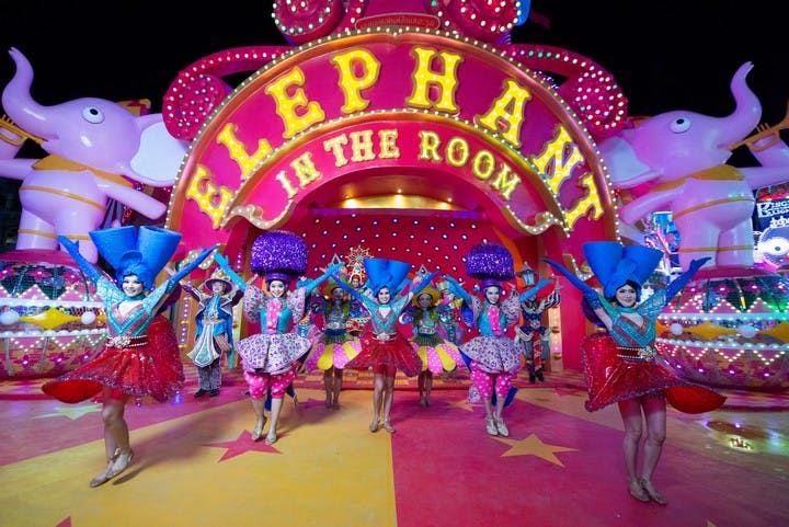 화려한 의상을 입은 공연자들은 분홍색 코끼리 동상을 뒤로 한 채 '방 안의 코끼리' 라고 적힌 커다란 표지판 앞에서 춤을 추고 있습니다.