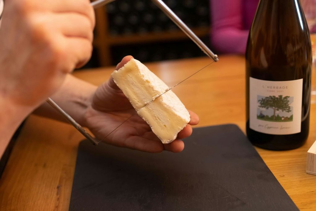 검은 도마 위에 와이어 치즈 커터로 치즈 조각을 썰고 병을 배경으로 한 손.