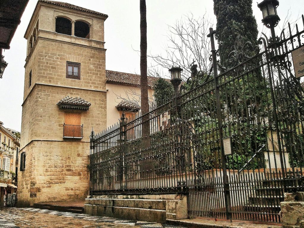 검은색의 화려한 철제 울타리, 자갈길, 높은 나무를 배경으로 막힌 창문이 있는 석조 건물입니다.