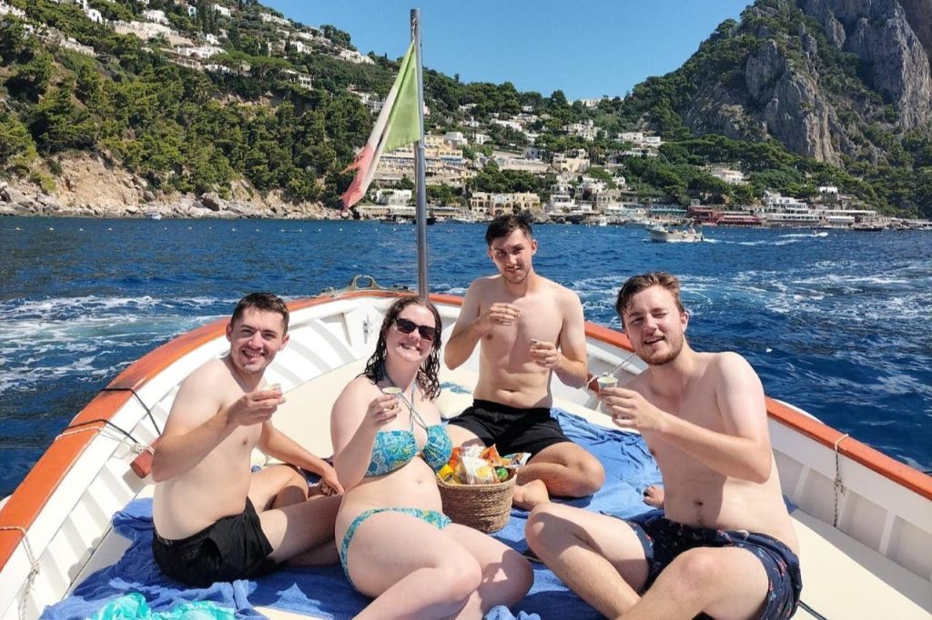 태양 아래 보트에 앉아 다과를 즐기고 있는 세 사람은 아름다운 해안선을 배경으로 아름다운 풍광을 자랑합니다.