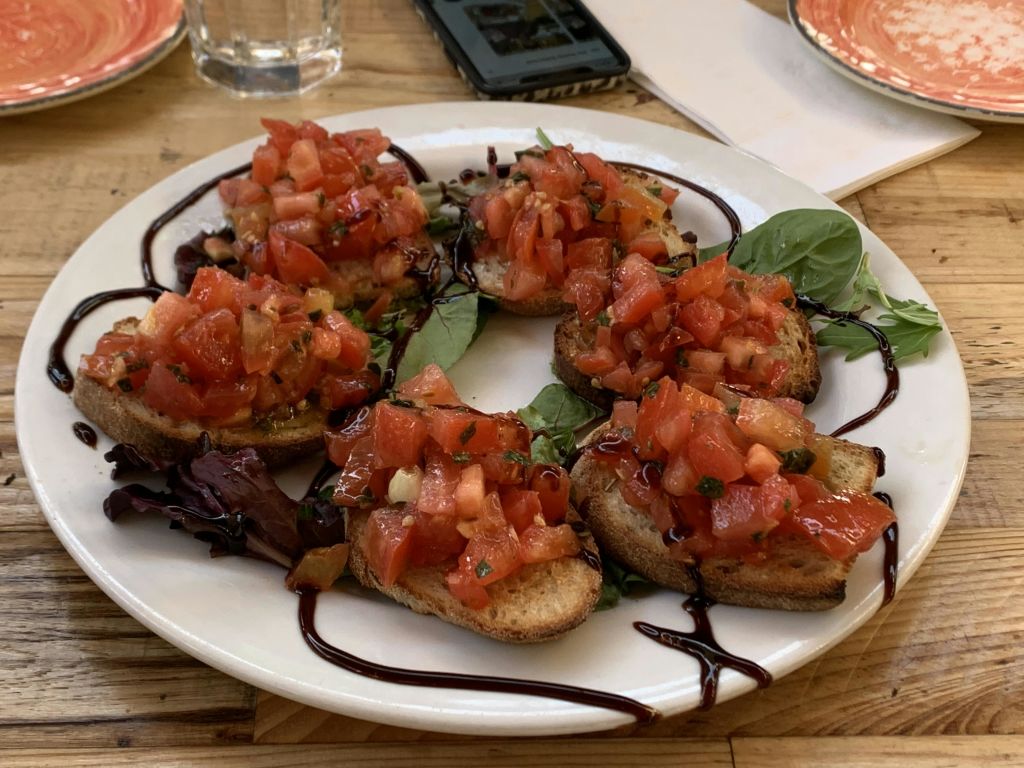 잘게 썬 토마토를 토핑하고 녹색으로 장식한 브루스케타 한 접시에 발사믹 유약을 뿌려줍니다. 배경에는 전화기와 유리가 있습니다.