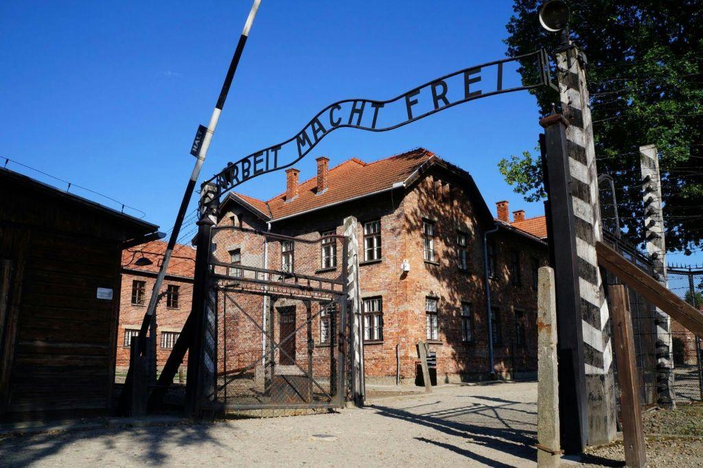 맑고 푸른 하늘 아래 벽돌 건물로 이어지는 "Arbeit Macht Frei" 표지판이 있는 아치형 철문.