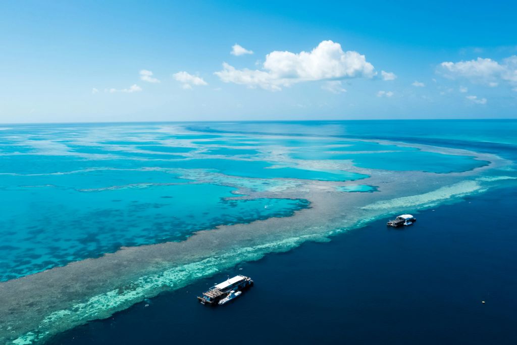 두 척의 보트가 산호초 근처에 정박해 있으며, 맑고 청록색의 물이 밝고 푸른 하늘 아래 구름 몇 개가 흩어져 있습니다.