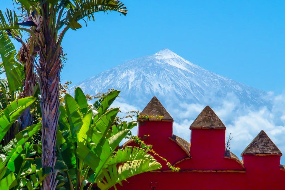 맑고 푸른 하늘 아래 녹색 열대 단풍으로 둘러싸인 뾰족한 지붕이 있는 붉은 건물 뒤의 눈 덮인 산.